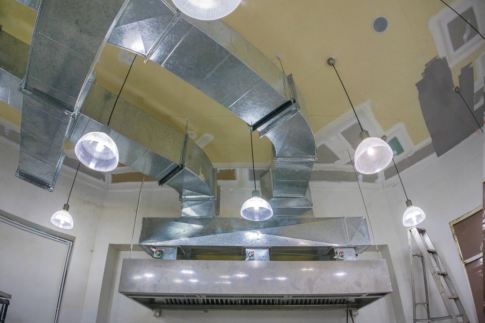 Industrial ceiling with large silver ductwork, hanging lights, and beige walls in a room under construction