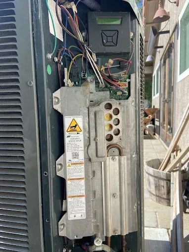 Open electrical panel with exposed wiring and warning label beside an outdoor unit
