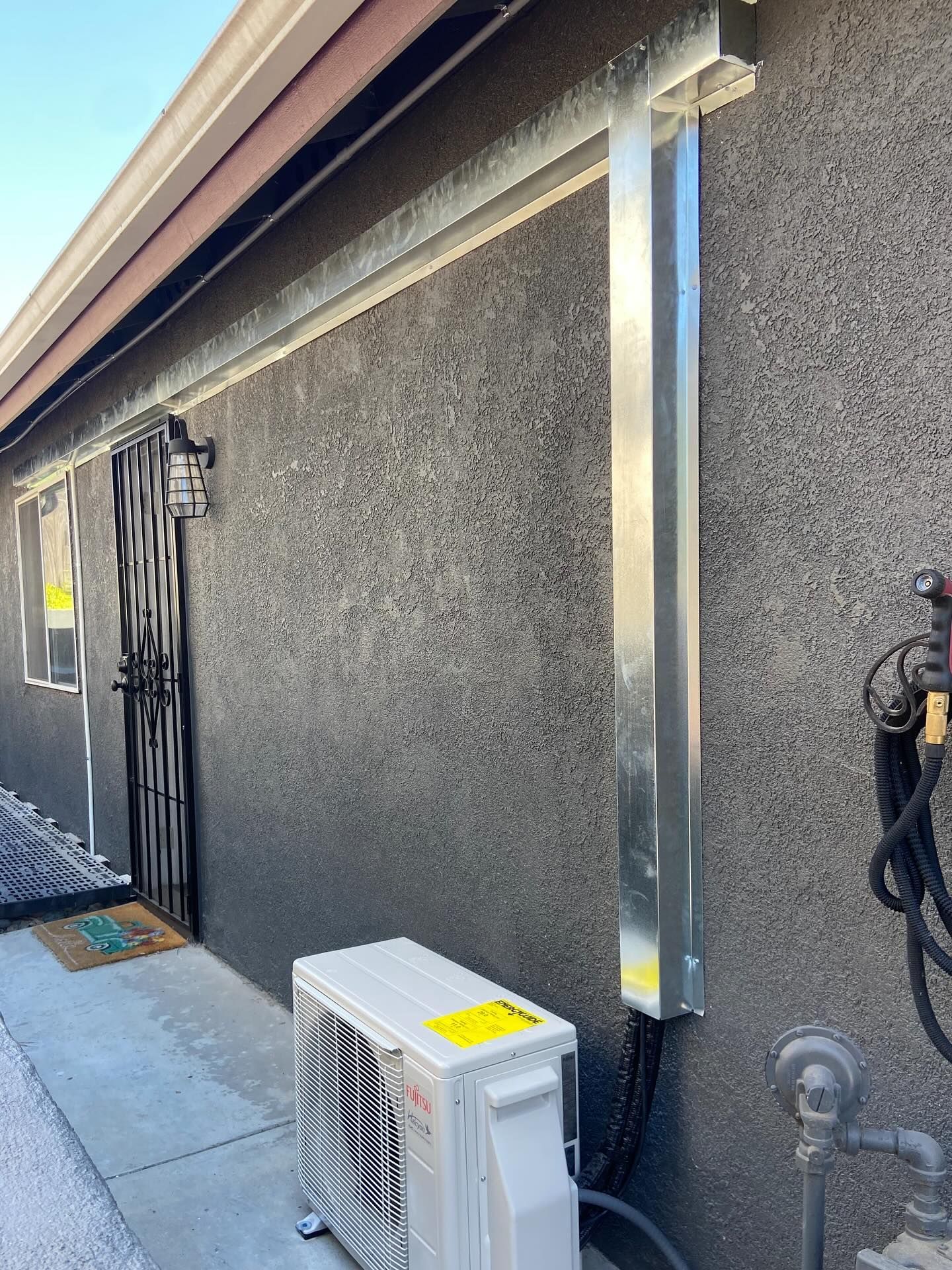 Exterior wall with mounted utility conduit and an air conditioner unit beside a concrete walkway.