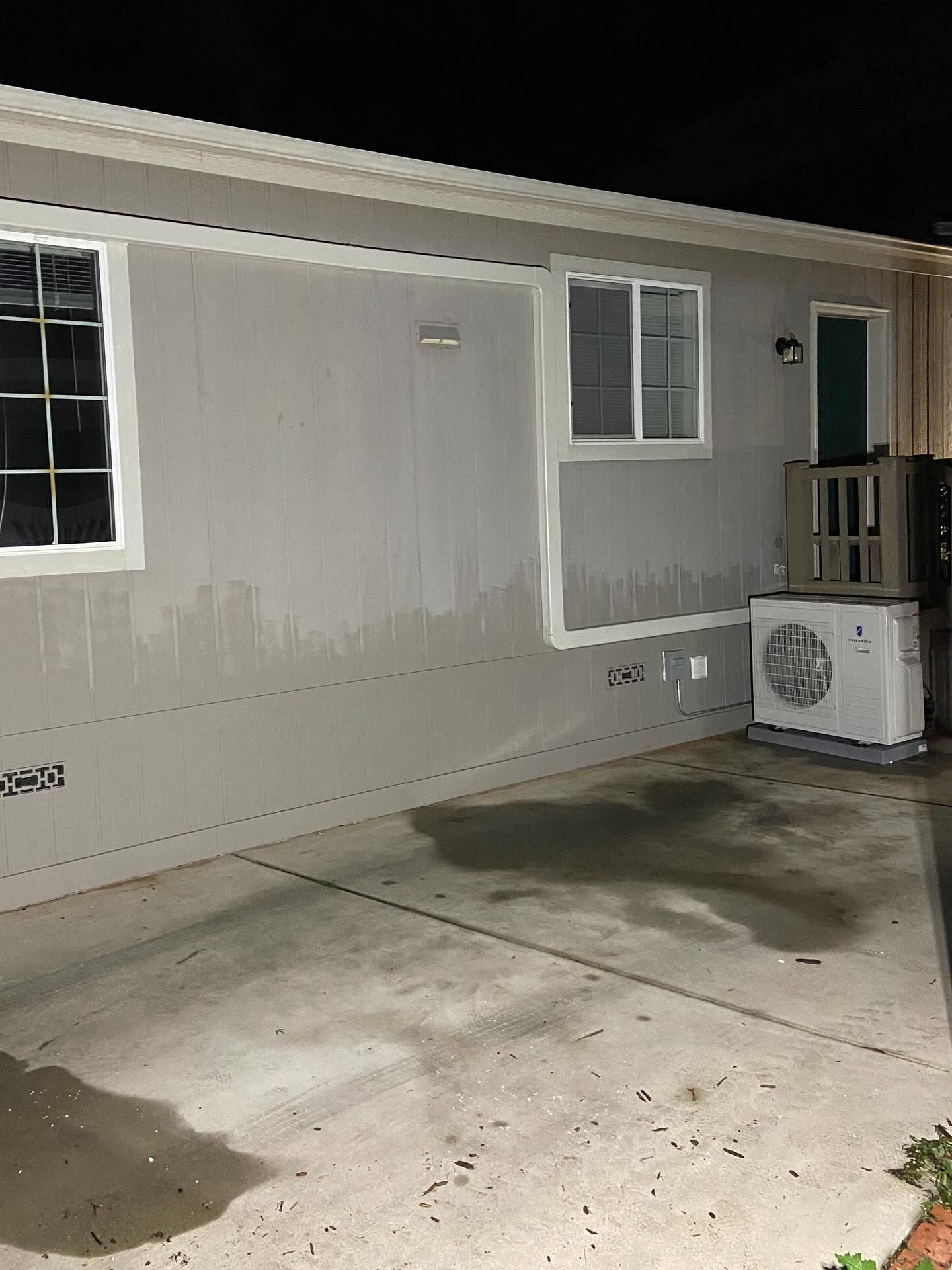 Mobile home exterior at night with concrete patio, wet stains, and an air conditioner unit by the door