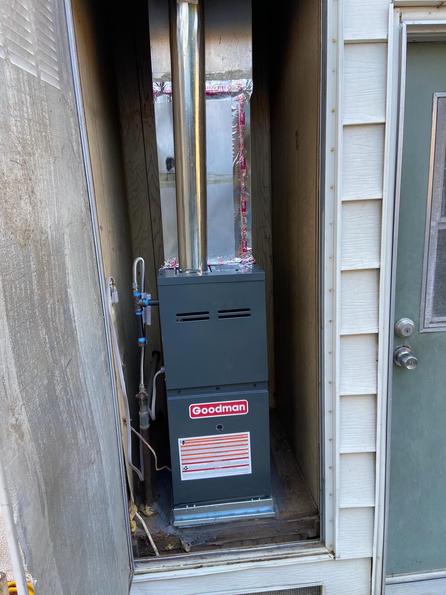 Gas furnace in a narrow outdoor utility alcove beside house siding and a metal door