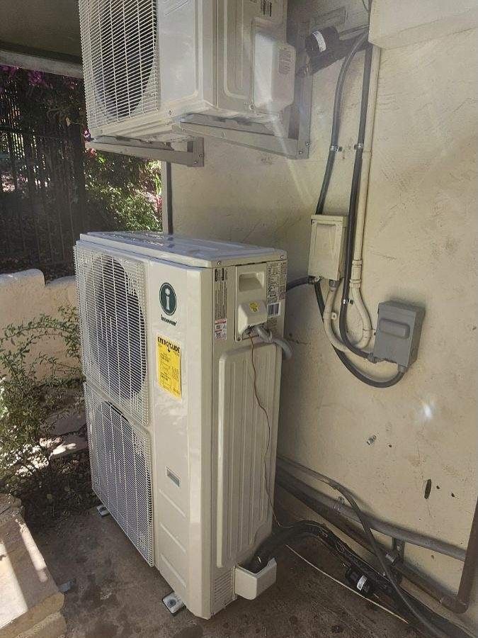 Two outdoor air conditioning units beside a wall, with electrical conduit and cables.