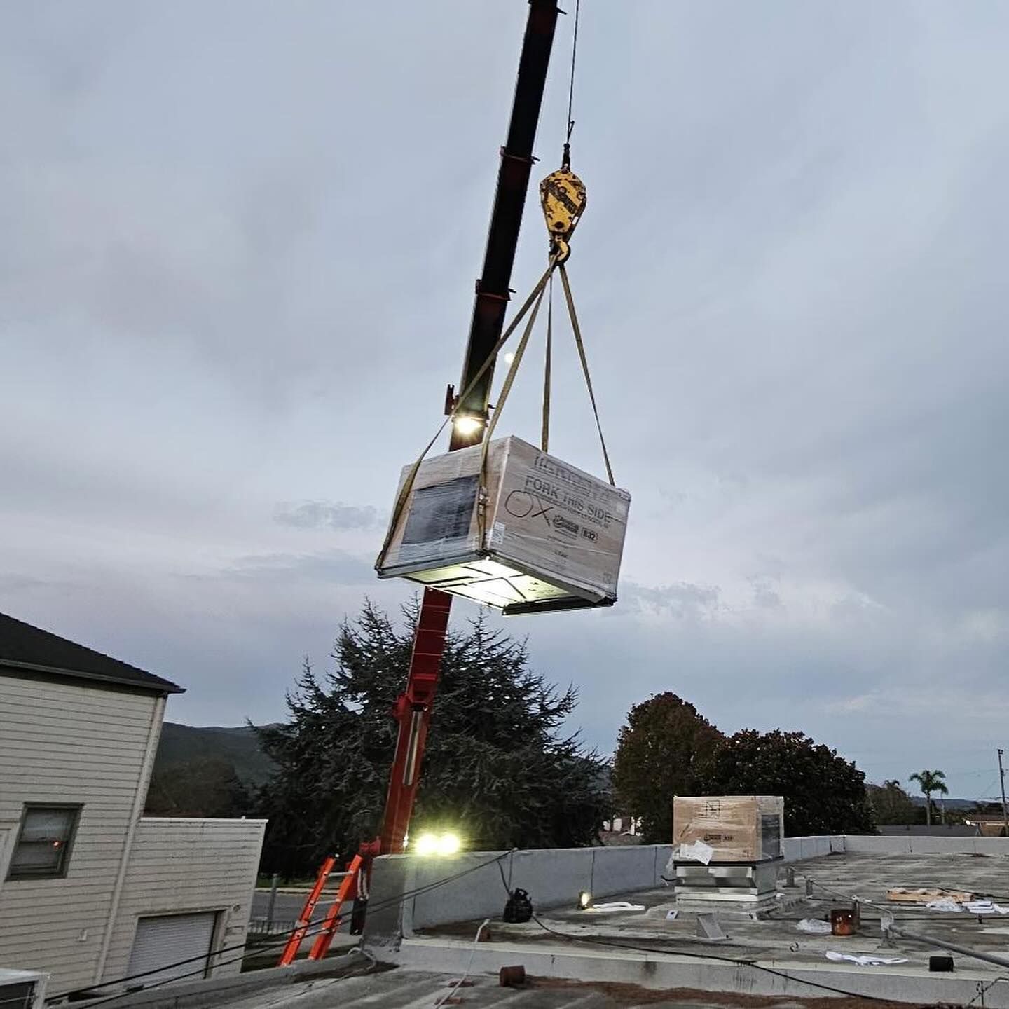 Crane lifting a wrapped HVAC unit above a flat rooftop at dusk