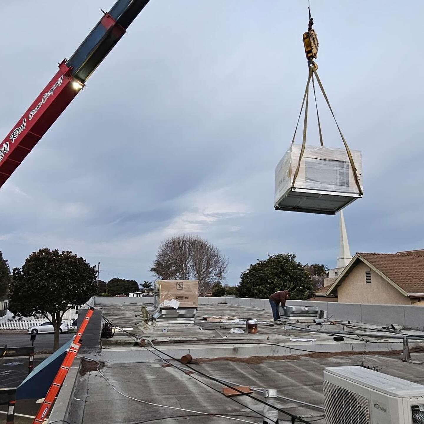 Crane lifts a bundled rooftop HVAC unit over a flat roof with workers below on an overcast day