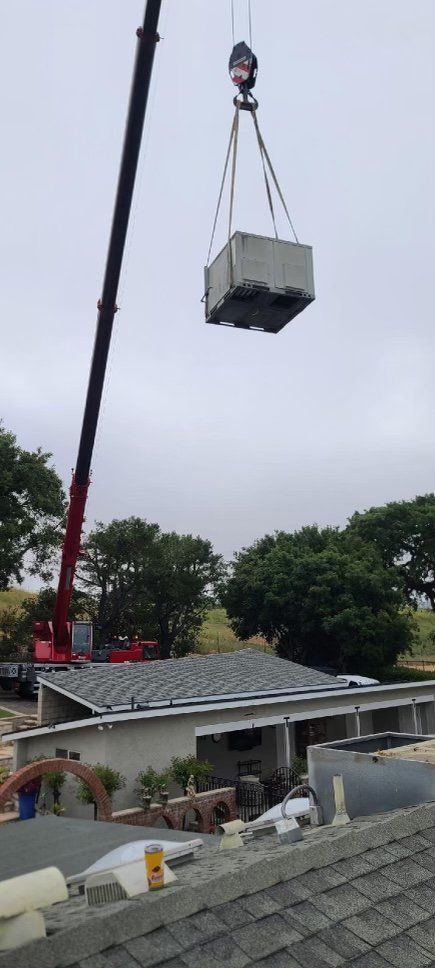 Crane lifting a concrete block over a residential roof beside trees and houses