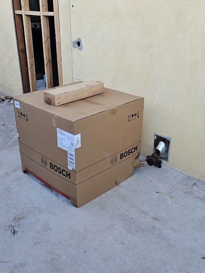 Two stacked cardboard boxes on a concrete floor beside a wall outlet and wooden frame opening.