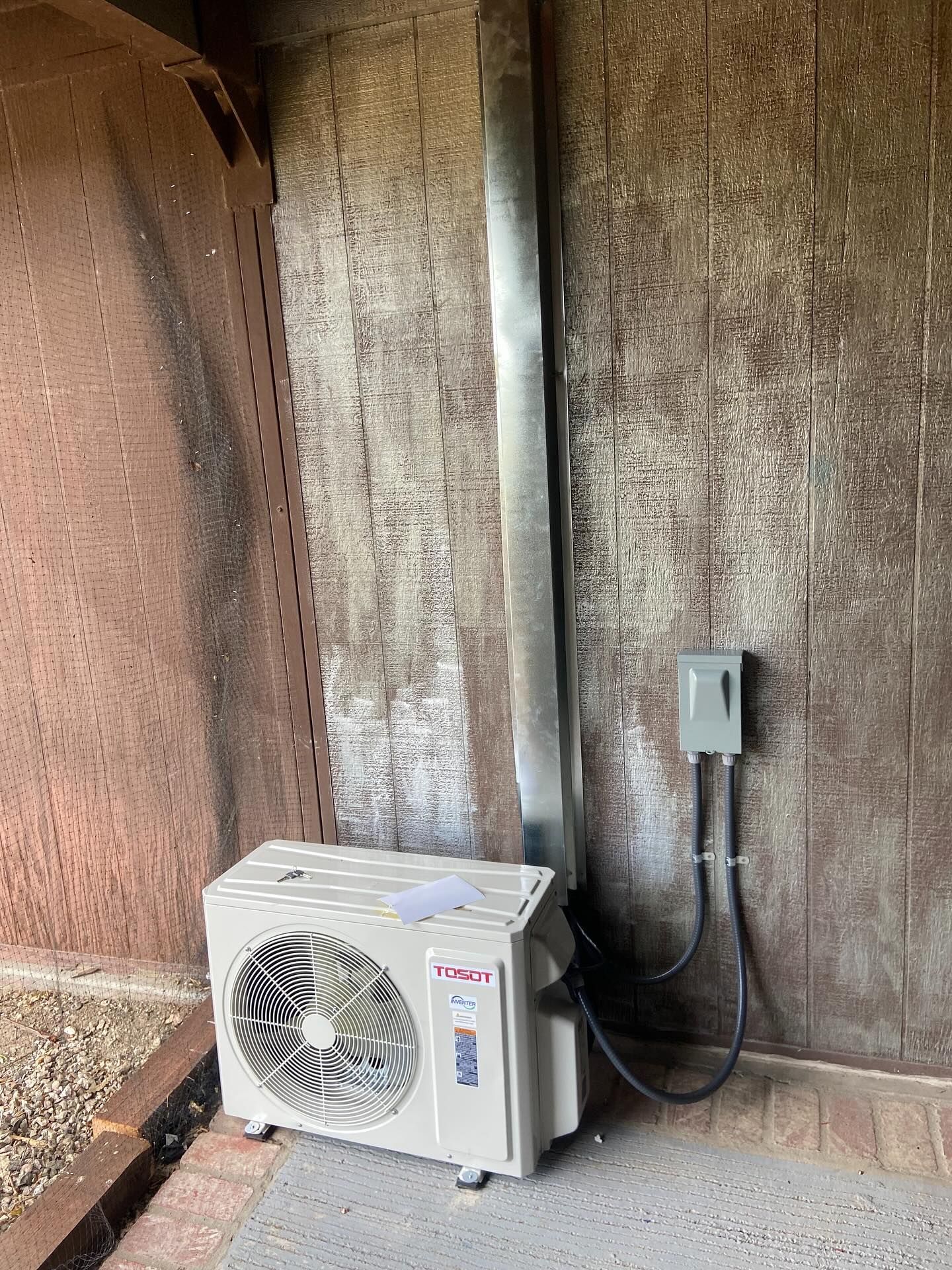 Outdoor heat pump unit beside a concrete wall with electrical conduit and outlet box
