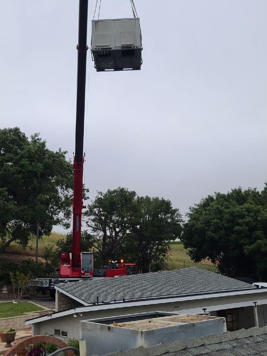 Crane lifting a gray utility box above a building roof with trees and an overcast sky in the background