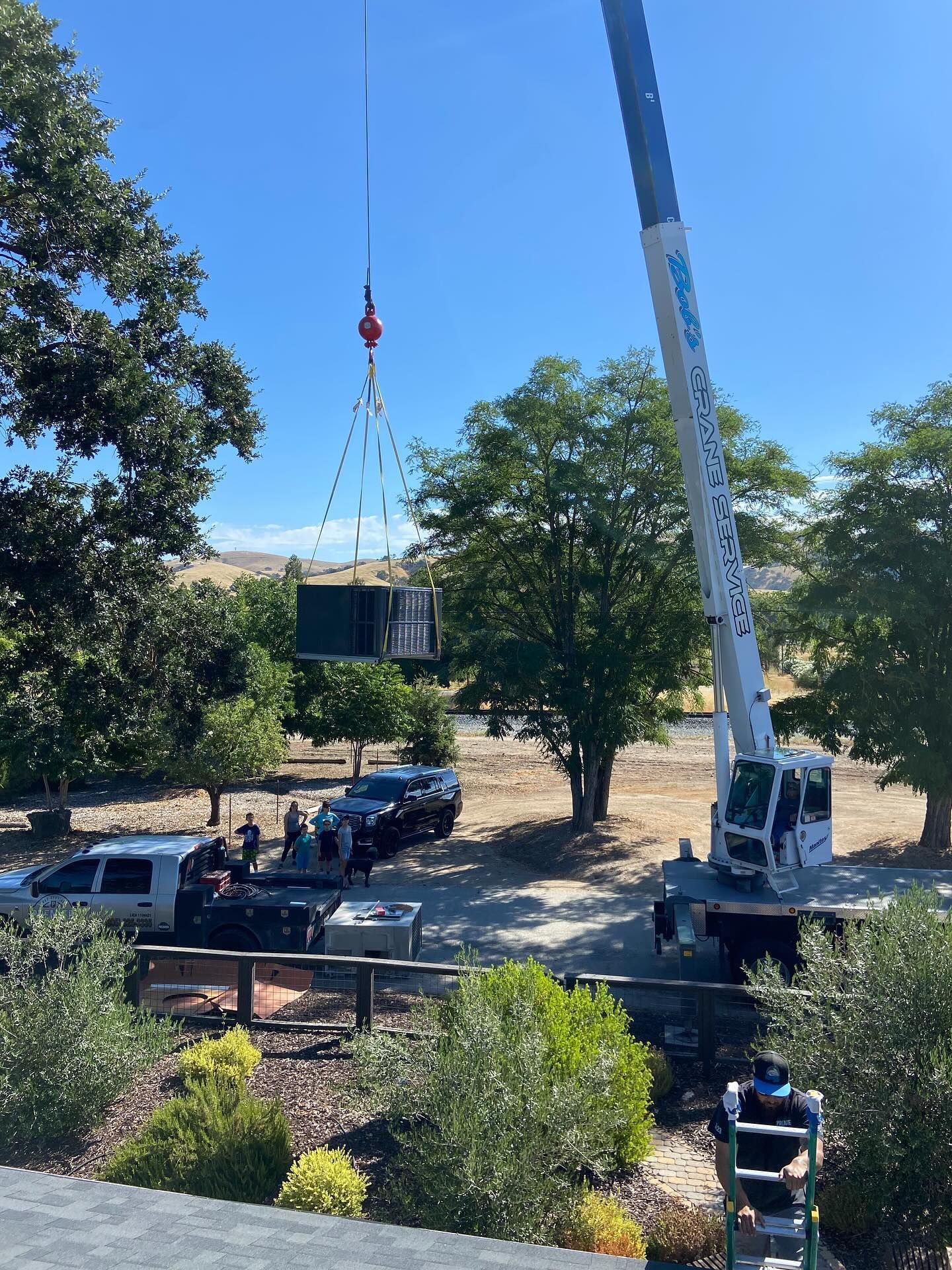 Crane lifting a large black object over a parking lot surrounded by trees and parked cars