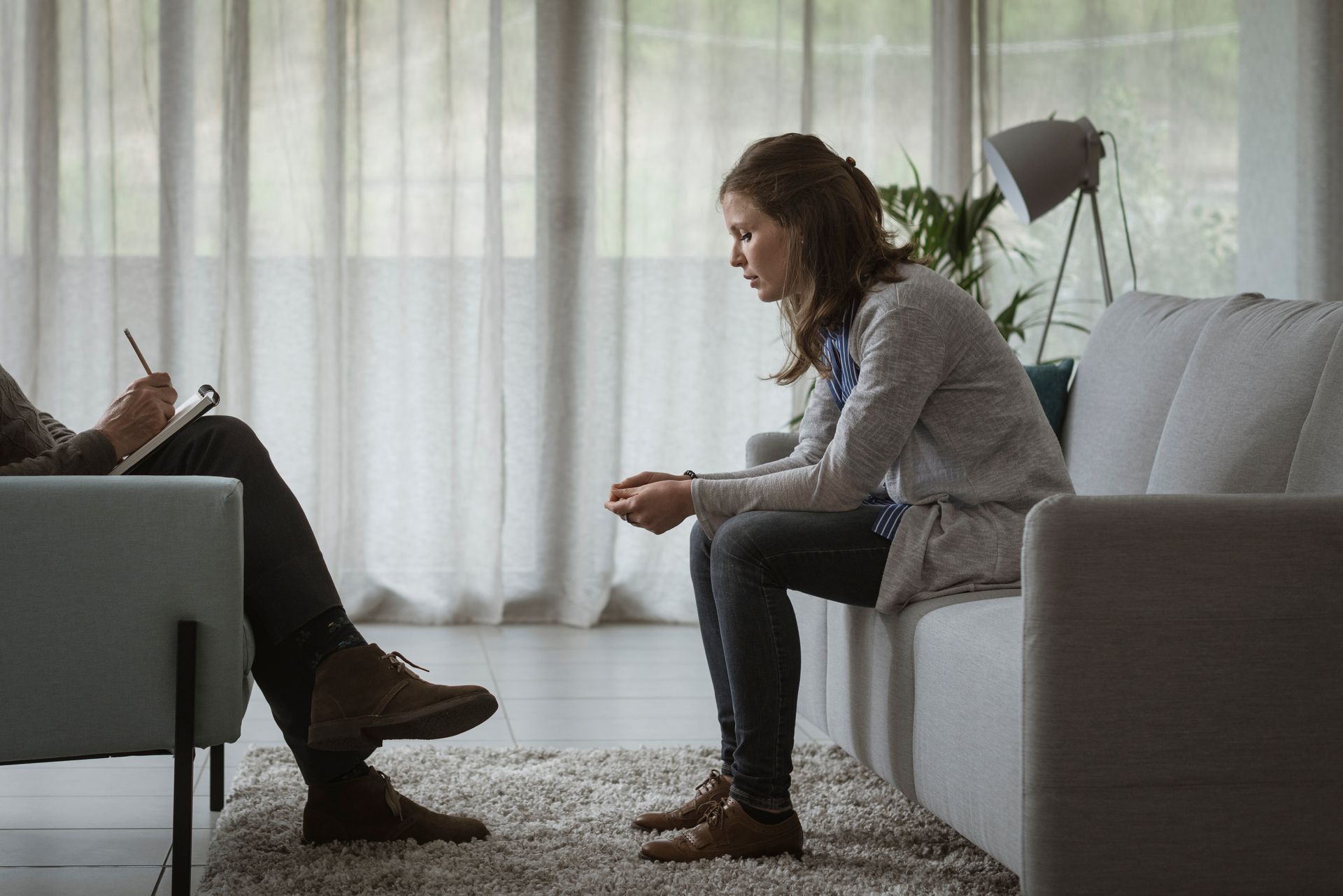Woman sits on sofa, leaning forward and talking in a bright living room with sheer curtains.