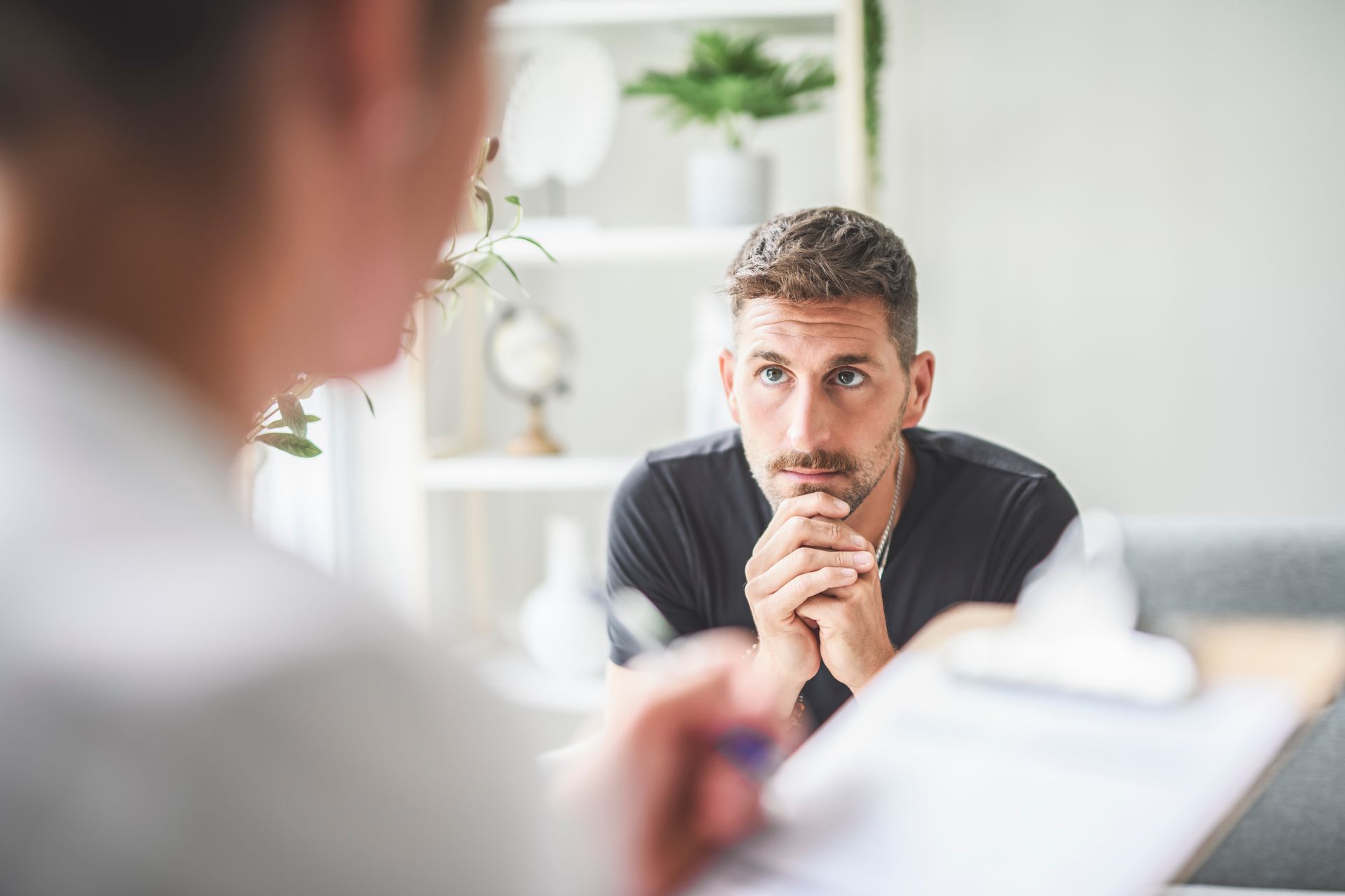 Man listening intently across a table in a bright office, hands clasped, with another person blurred in foreground
