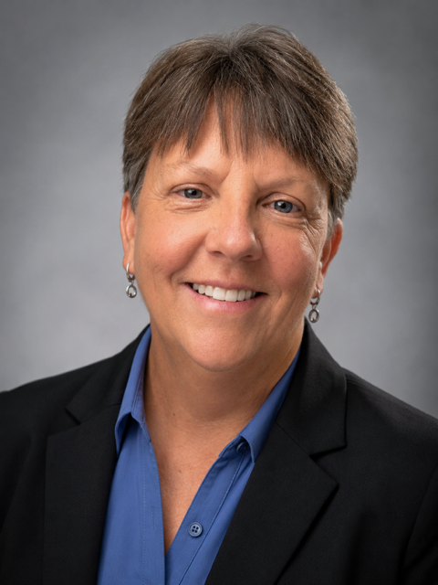 Smiling person in a dark blazer and blue shirt against a gray background