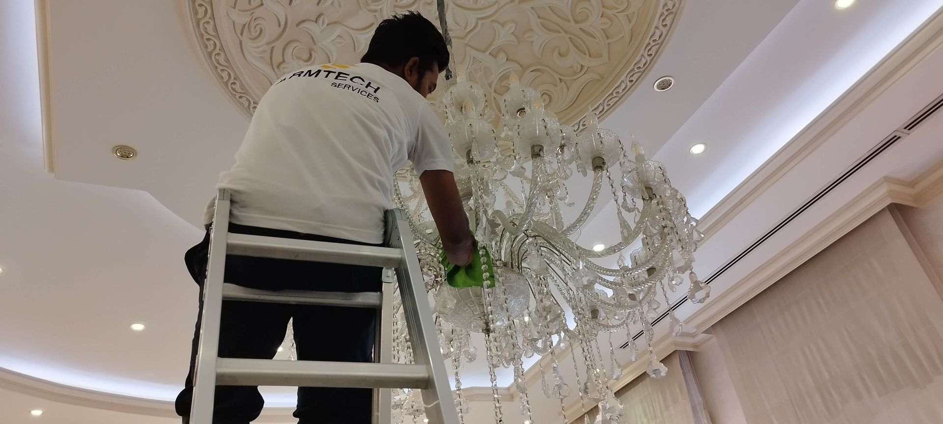 A man is standing on a ladder cleaning a chandelier.