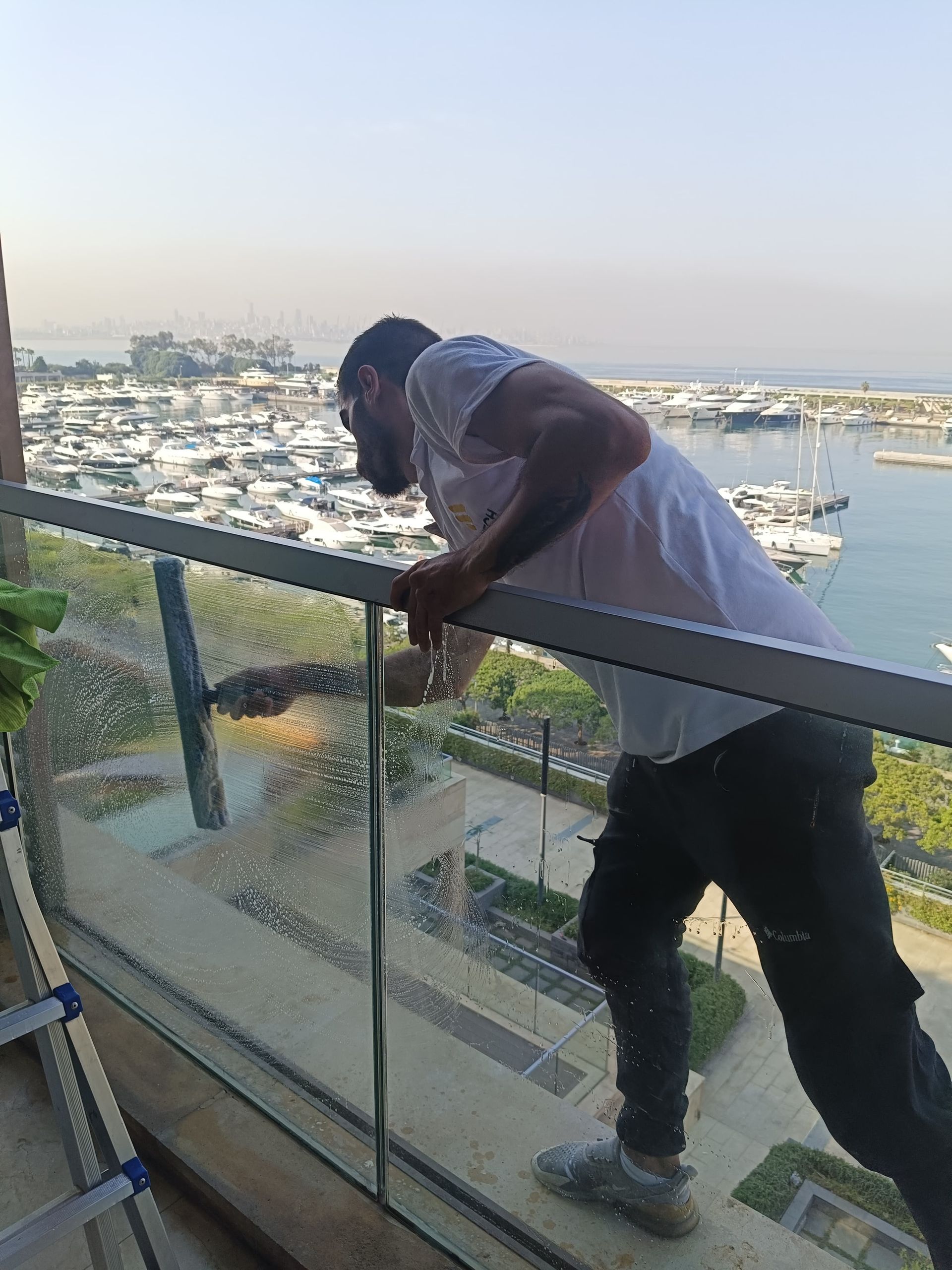 A man is cleaning a glass railing on a balcony overlooking a marina.