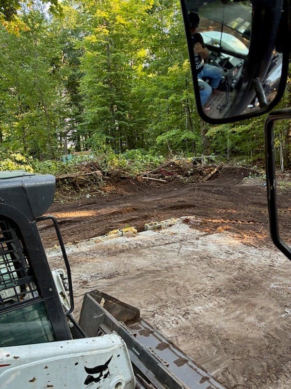 View from a skid steer over a cleared dirt area beside a wooded path, with a rearview mirror visible.