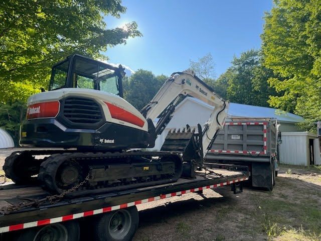 Mini excavator on a flatbed trailer outdoors beside a shed and trees.