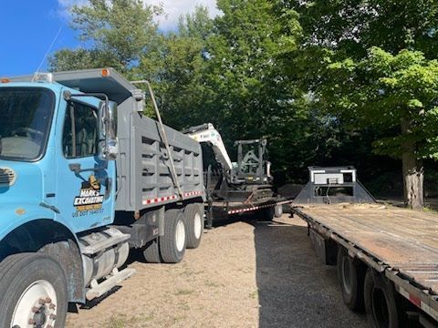 Blue dump truck hauling heavy equipment beside a flatbed trailer on a gravel lot, with trees in the background