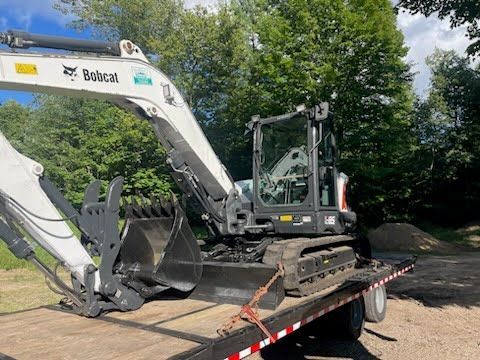 White excavator loaded on a flatbed trailer in a wooded area.