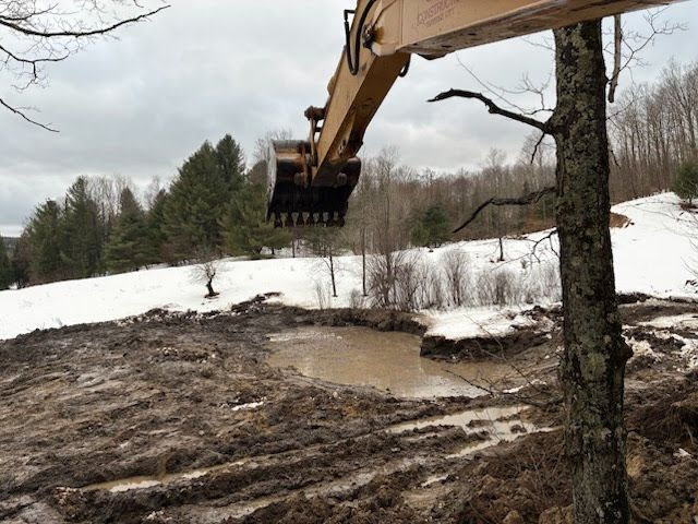 Excavator digging muddy ground near a snowy roadside and leafless trees