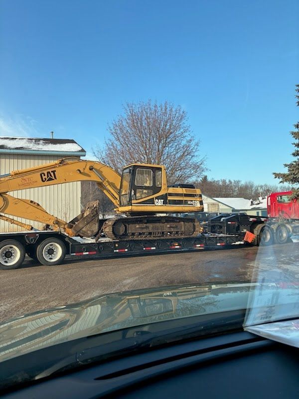 Yellow excavator on a flatbed trailer on a road, viewed from inside a car under a blue sky