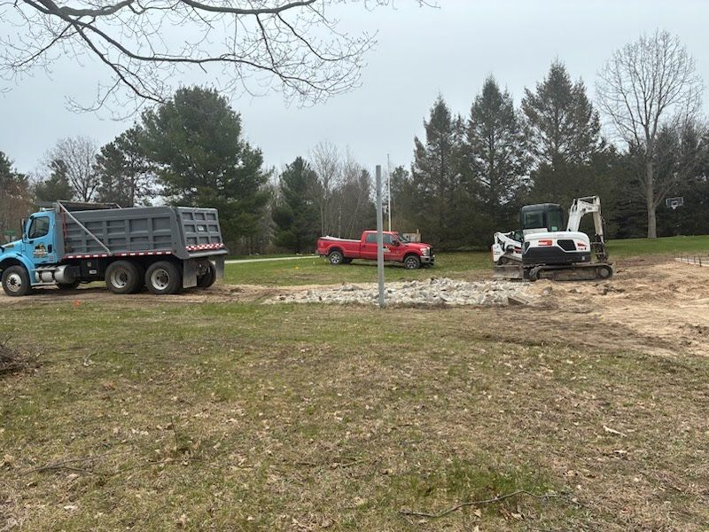 Blue dump truck, red pickup, and white backhoe parked around a gravel area with trees in the background