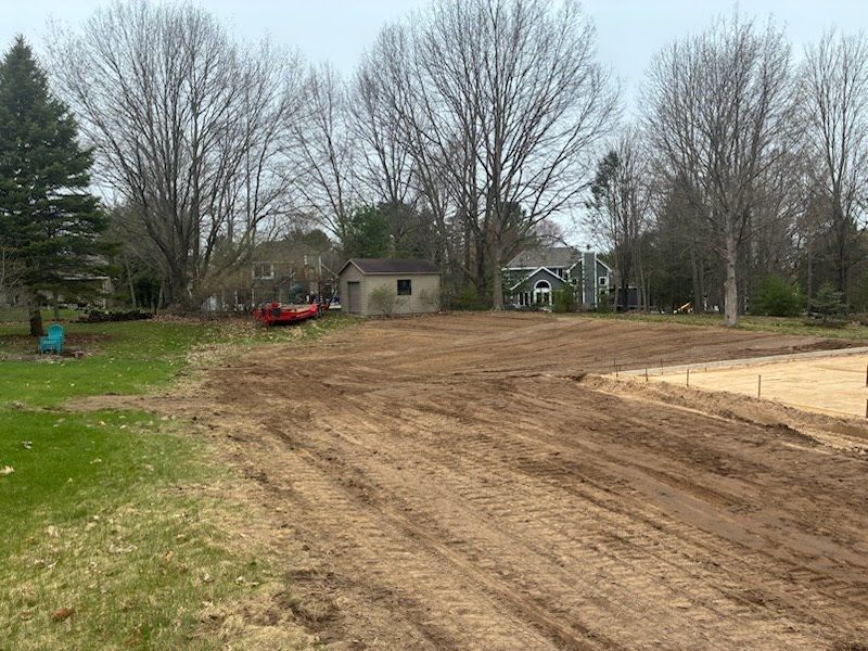Cleared dirt lot beside grassy yard with bare trees, a small shed, and a red vehicle in the background