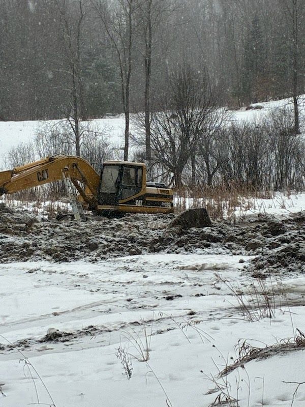 Yellow excavator working in a snowy muddy field with bare trees in the background