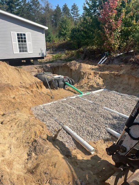 Trench excavation beside a house with gravel, exposed pipes, and green piping in a dirt construction site