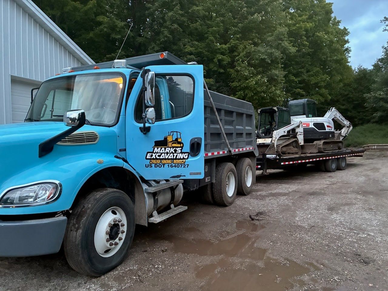 Blue dump truck hauling a flatbed trailer with equipment on a muddy roadside
