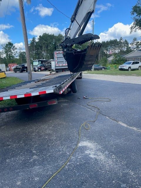Excavator loading onto a flatbed truck in a parking lot, with broken pavement and a chain nearby