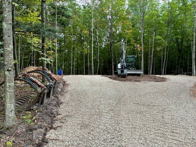 Gravel driveway in a wooded area with a small excavator and stacked logs on the left