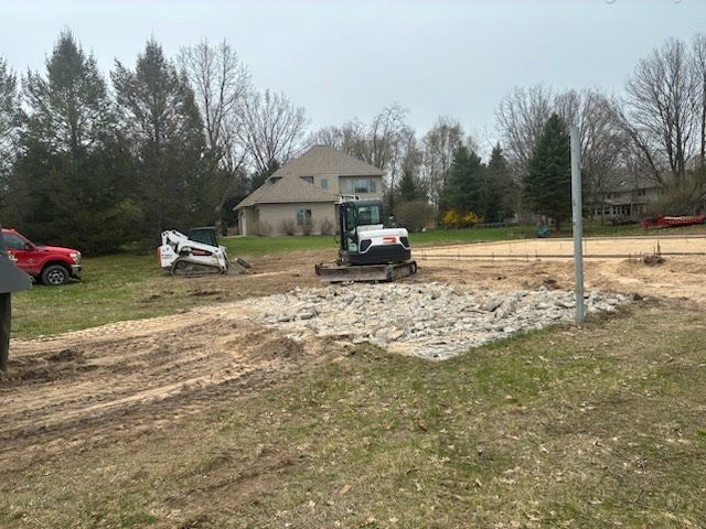 Small skid steer on a dirt lot near a house, with a red truck and trees in the background.