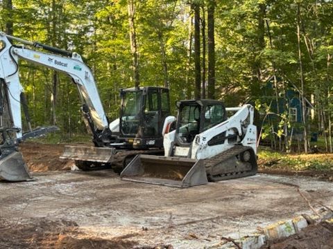 Two construction machines, an excavator and a skid steer, parked on a gravel site near trees.