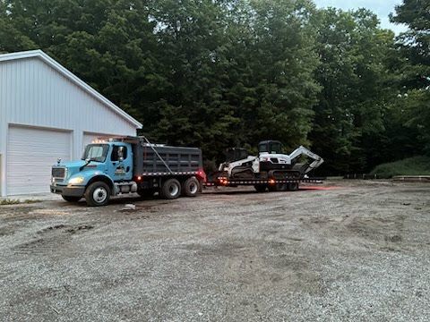 Blue dump truck towing a flatbed trailer beside a white garage in a gravel lot at dusk