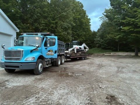 Blue tow truck hauling a white jet ski on a flatbed in a gravel lot beside a building and trees.