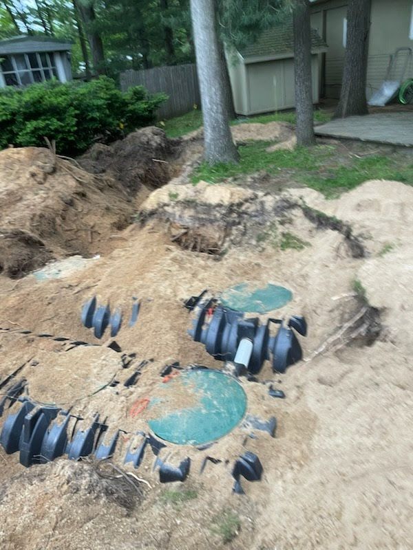 Underground utility pit with exposed blue and black pipes in a dirt yard near trees and a house