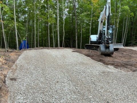 Construction site with excavator beside a large gravel mound in a wooded area