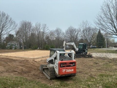 White skid steer on a dirt lot with excavator and trees in the background