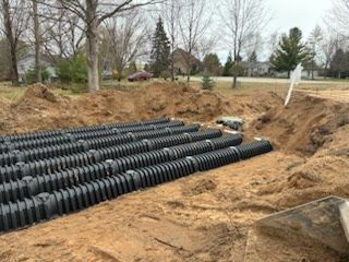 Rows of black corrugated drainage pipes stacked in a dirt excavation site with bare trees nearby