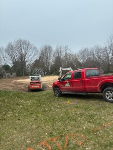 Red pickup truck and orange skid steer in a grassy yard under a cloudy sky