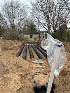 Black drainage pipes laid in a sandy trench beside a plastic-covered post and bare trees