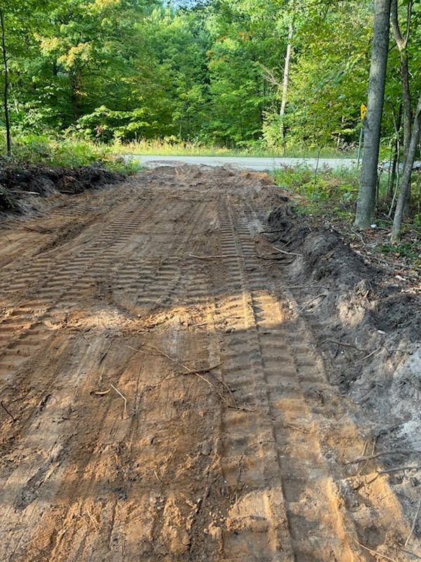 Dirt road under construction with tire tracks, bordered by green trees and brush.