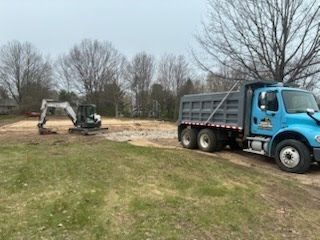 Blue dump truck and excavator working in a muddy field with bare trees nearby