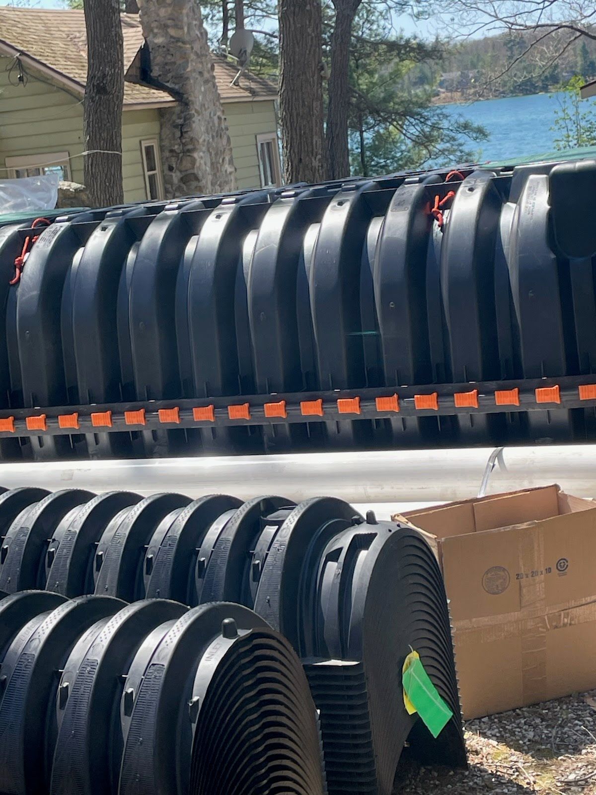 Stacked black ribbed plastic pipes outdoors near a lake, with trees and boxes in the background.