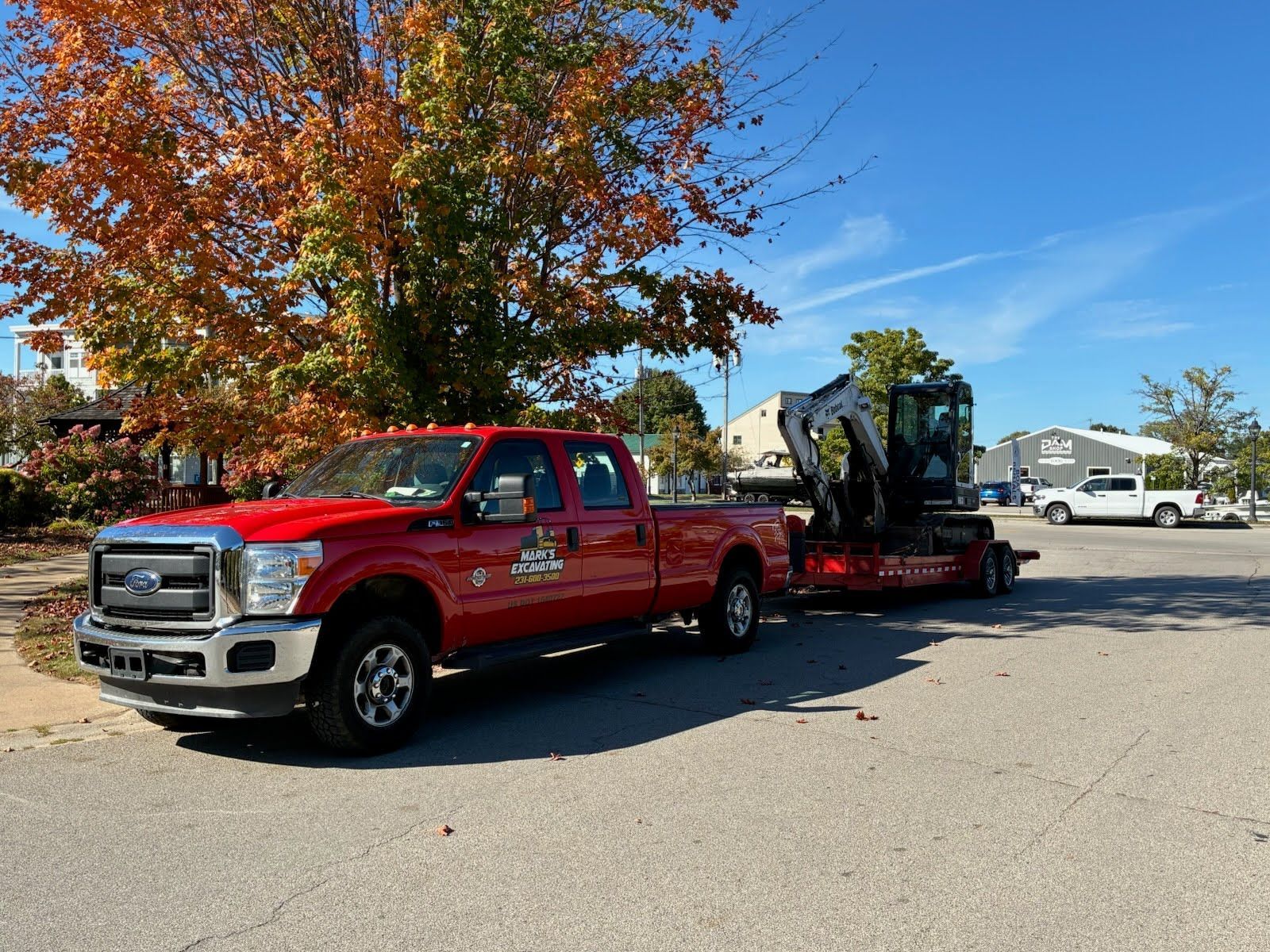 Red pickup truck towing a trailer with a small excavator on a suburban street in fall.