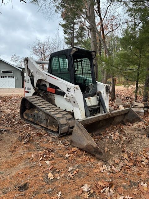 White skid-steer loader with bucket parked on dirt and leaves in a wooded area