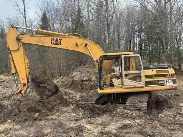 Yellow CAT excavator digging in a muddy wooded area, with an operator in the cab.
