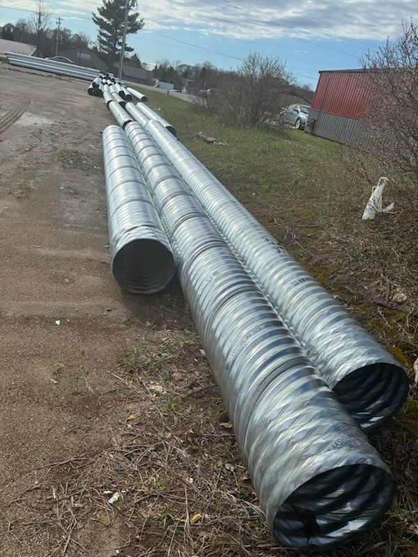 Several large silver corrugated metal pipes lying on a dirt lot near a red building