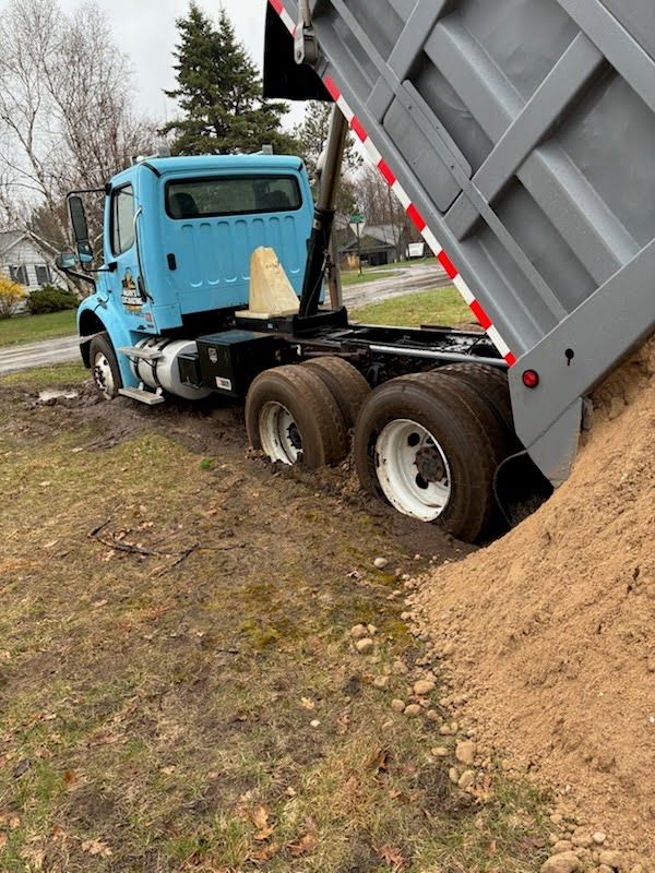 Blue dump truck tipped on muddy grass, unloading a large pile of sand beside a roadside.