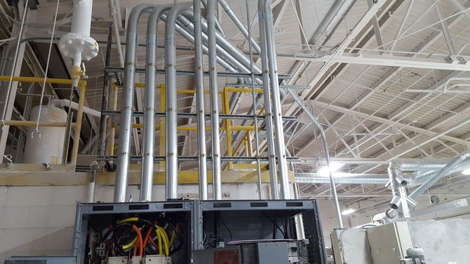 Industrial ceiling with exposed silver pipes and yellow conduit above an open electrical panel.
